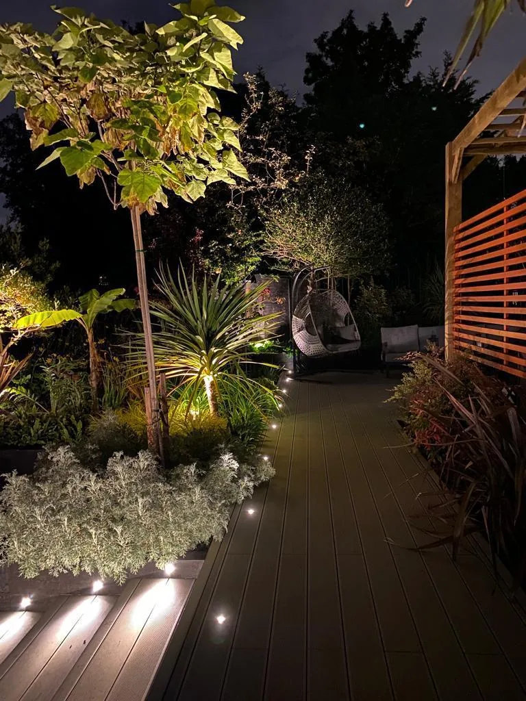 Illuminated garden walk with uplit mature plants and a red painted trellis screen
